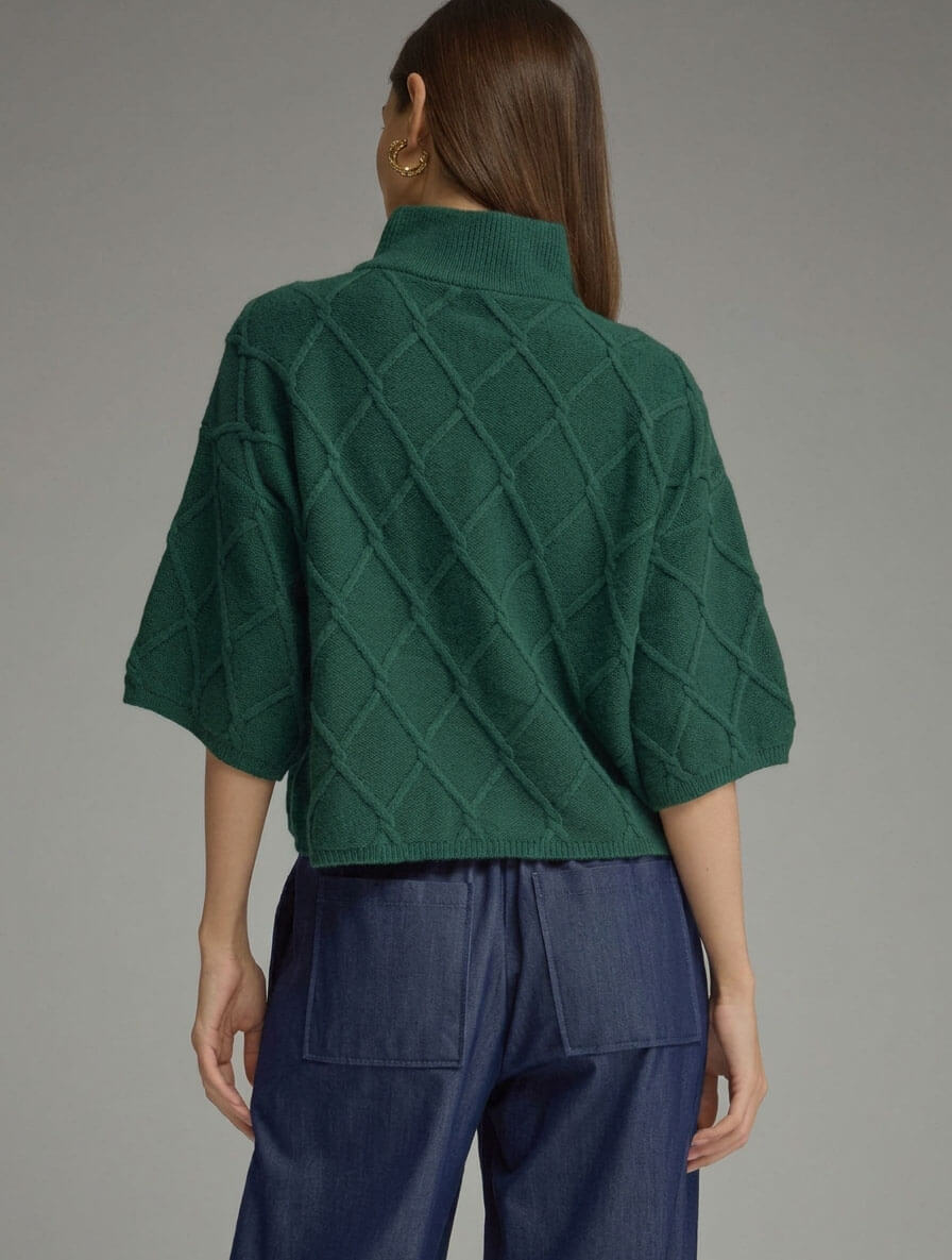 A woman shown from the back wearing a dark green knit sweater with a diamond cable pattern and a mock neck, paired with dark blue pants, against a solid gray studio background.